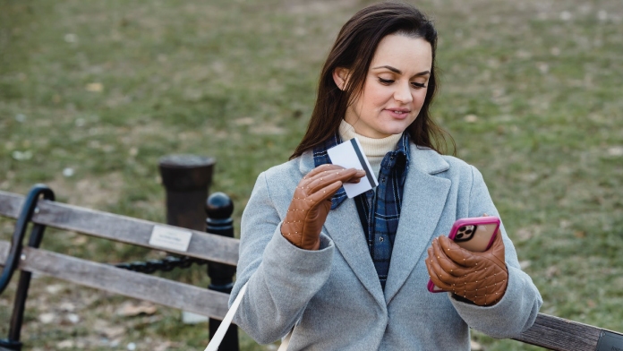 mulher caminhando num jardim com luvas de inverno e de cartão de crédito na mão enquanto olha para o smartphone