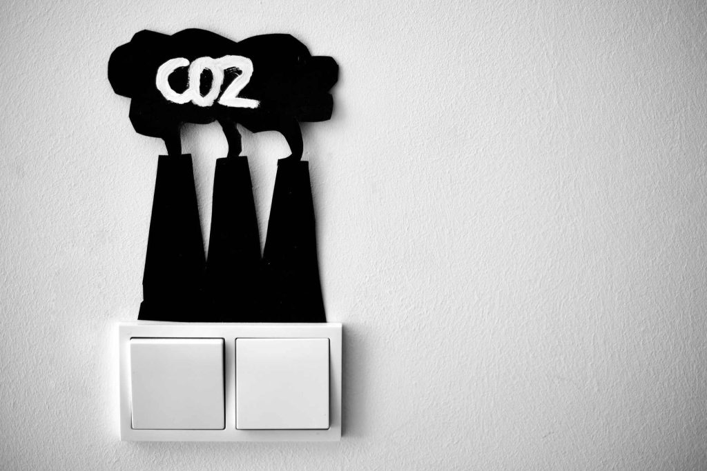 cartão colado na parede, por cima de interruptores elétricos. O catão, de cor preta, está recortado com a forma de nuvem negra, com a palavra "CO2" escrita a branco sobre chaminés industriais.