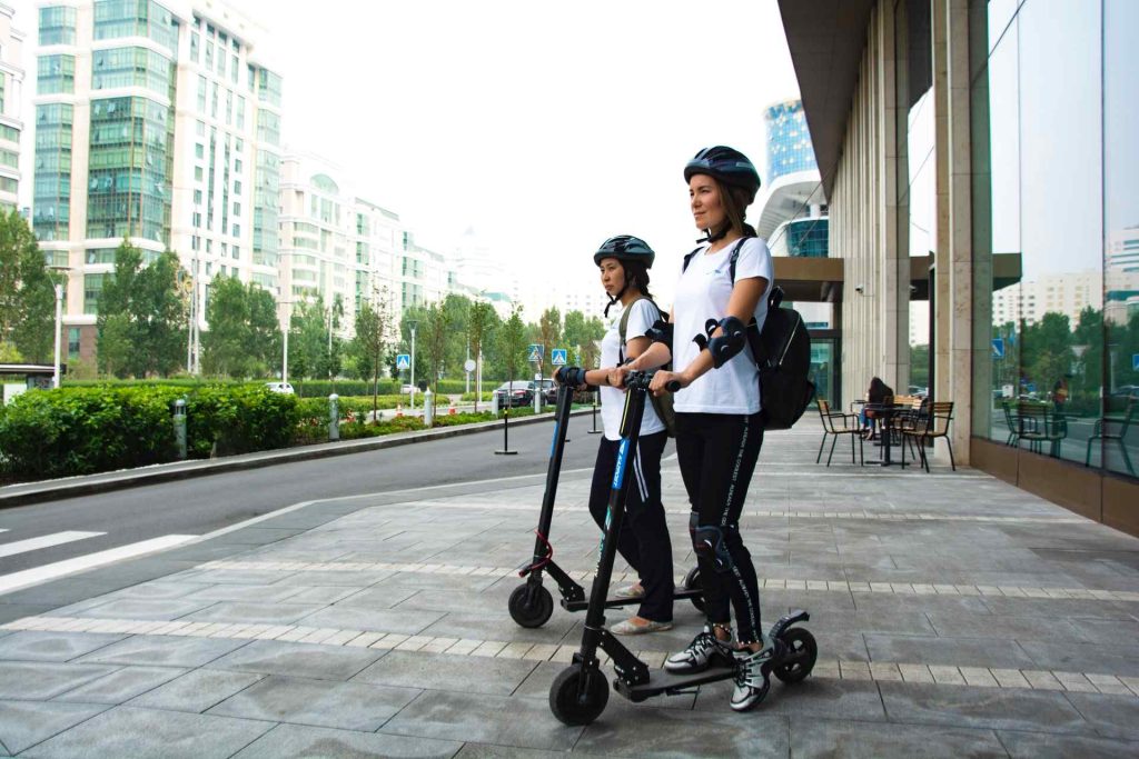 Mulheres com trotinetes no passieo de uma cidade moderna.