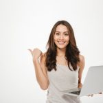 amazing-businesswoman-holding-silver-laptop-and-pointing-finger-sideways-with-smile-over-white-wall